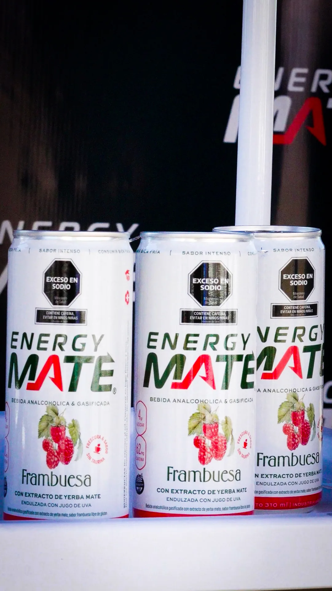 Tres latas de Energy Mate frambuesa alineadas frente al cartel de la marca